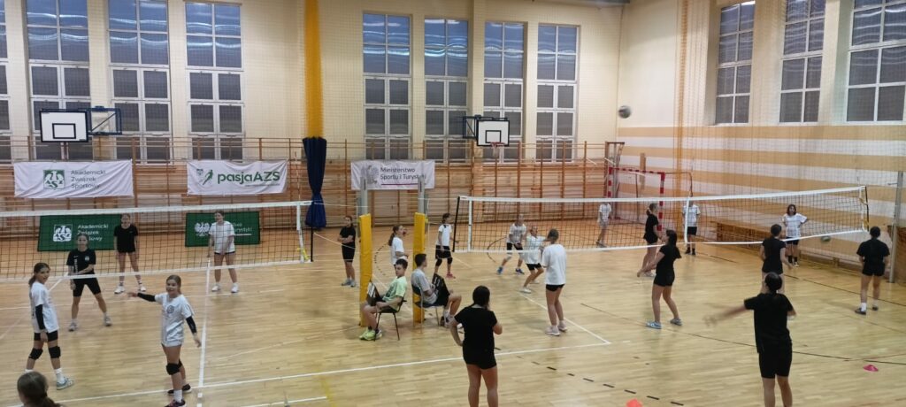 Zdjęcie przedstawia zajęcia sportowe w sali gimnastycznej, gdzie uczestnicy grają w siatkówkę. Widać dwie rozstawione siatki i kilka drużyn równocześnie rozgrywających mecze. Uczestnicy są w różnym wieku, ubrani w czarne lub białe stroje sportowe. Część osób przygotowuje się do odbioru piłki, inni obserwują grę z boku. W tle znajdują się banery z napisami: “Akademicki Związek Sportowy”, “Ministerstwo Sportu i Turystyki” oraz “pasjaAZS”, co sugeruje, że wydarzenie jest częścią programu promującego aktywność fizyczną. Duże okna sali gimnastycznej wpuszczają światło, a wystrój hali wskazuje na typowe miejsce szkolnych zajęć sportowych. Atmosfera na zdjęciu wydaje się dynamiczna, pełna zaangażowania i ruchu.
