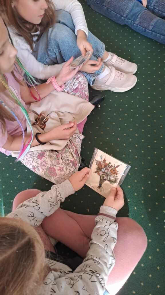 Na zdjęciu trzy dziewczynki ubrane w kolorowe ubrania siedzą na zielonym dywanie. Dziewczynka na pierwszym planie siedzi w siadzie skrzyżnym do nas tyłem. W ręku trzyma kolorową pocztówkę w brązowych barwach. Na pocztówce zabytki Olsztyna ułożone w bukiet.