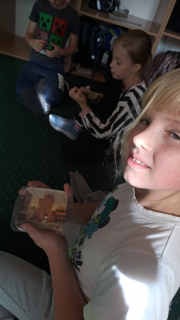 Na zdjęciu siedzi trzech chłopców. Siedzą obok siebie na zielonym dywanie. Blondyn na pierwszym planie trzyma w ręku magnes z widokiem olsztyńskiej Wysokiej Bramy. Na dalszym planie dziewczynka w koszuli w pionowe pasy także coś trzyma w dłoniach. Zdjęcie jest ciemne.