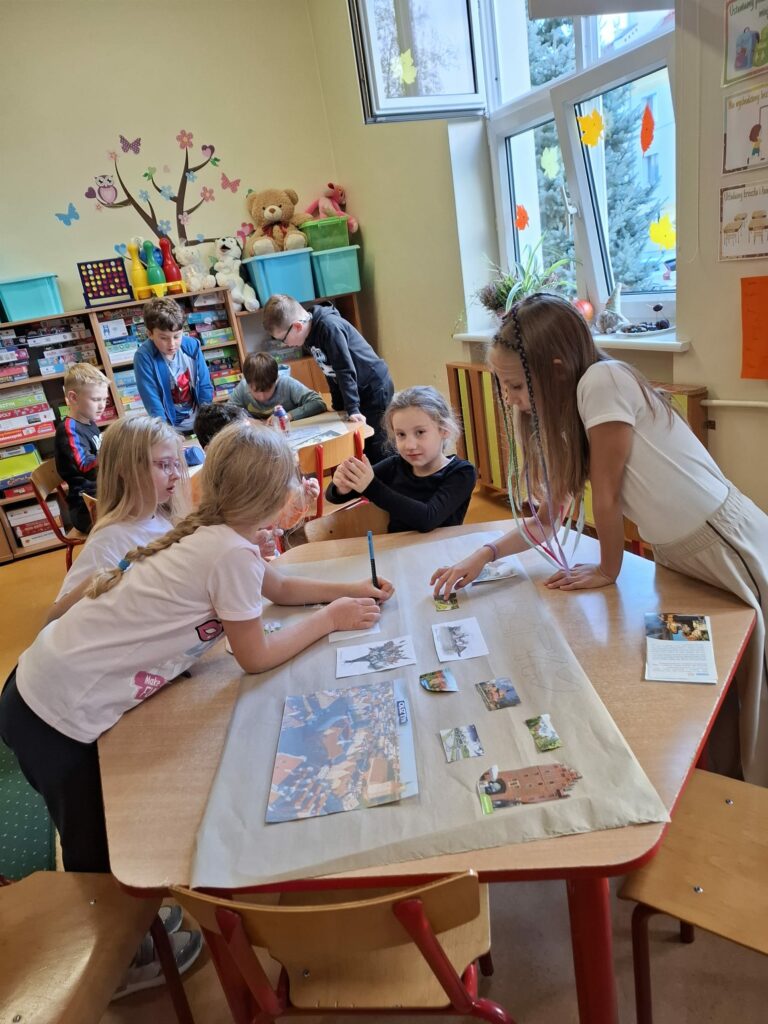 Na zdjęciu dwie dziewczynki ubrane w białe koszulki i jedna ubrana w czarną koszulkę pochylają się nad brązowym stołem. Na środku stołu leży szary, duży papier, na którym dziewczynki naklejają zdjęcia , robiąc pracę plastyczną. Na drugim planie widać okno oraz chłopców robiących swoją pracę plastyczną. Ściana jest koloru kremowego.