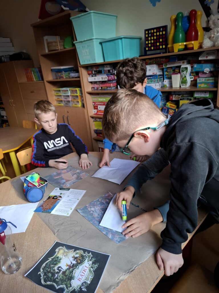 Na zdjęciu dwóch chłopców pochyla się nad brązowym stołem. Na środku stołu leży szary , duży papier na którym chłopcy naklejają zdjęcia , robiąc pracę plastyczną. Na stole leżą kartki z kolorowymi zdjęciami. Na drugim planie widać szafki z kolorowymi zabawkami, seledynowymi plastikowymi pudełkami.