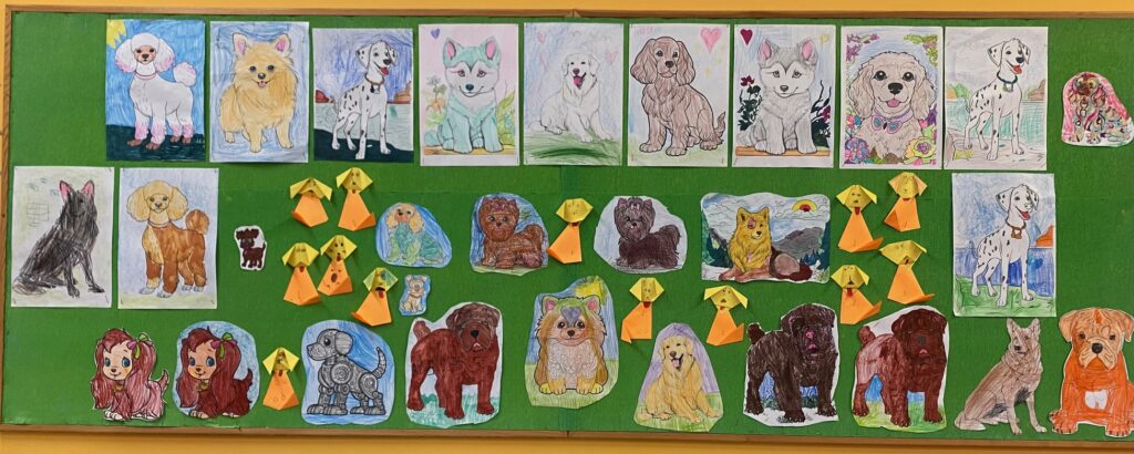 Tablica z pracami plastycznymi dzieci. Pokolorowane wizerunki psów oraz pieski origami. Na zielonym tle.