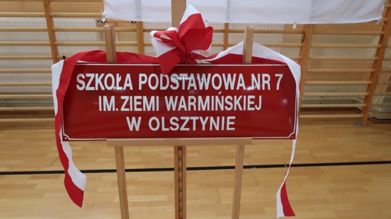 Na zdjęciu widzimy tablicę informacyjną z napisem: "Szkoła Podstawowa nr 7 im. Ziemi Warmińskiej w Olsztynie". Tablica ma czerwone tło i białe litery, a na jej górze widnieje biało-czerwona kokarda, symbolizująca oficjalną uroczystość, nadania szkole jej patrona. Tablica jest umieszczona na drewnianym stojaku, a w tle widać salę gimnastyczną z drewnianymi drabinkami na ścianie. Całość sugeruje uroczysty charakter wydarzenia odbywającego się w budynku szkoły.