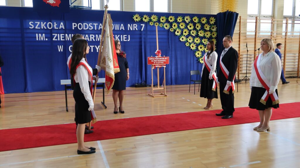 Na zdjęciu widać uroczystość szkolną, prawdopodobnie w Szkole Podstawowej nr 7 im. Ziemi Warmińskiej w Olsztynie, co sugeruje napis na tle w tle. Uczestnicy ceremonii stoją na czerwonym dywanie, ubrani w eleganckie stroje galowe, a niektórzy z nich noszą biało-czerwone szarfy. Jedna z osób trzyma sztandar szkolny z napisem „Szkoła Podstawowa nr 7” oraz herbem. Tło jest udekorowane granatowym materiałem z napisem i motywem słoneczników. Uroczystość odbywa się na sali gimnastycznej, co można wywnioskować po parkiecie i drabinkach gimnastycznych widocznych z tyłu. Atmosfera wskazuje na podniosły charakter wydarzenia, być może ślubowanie, nadanie imienia szkole lub inna ważna okazja.