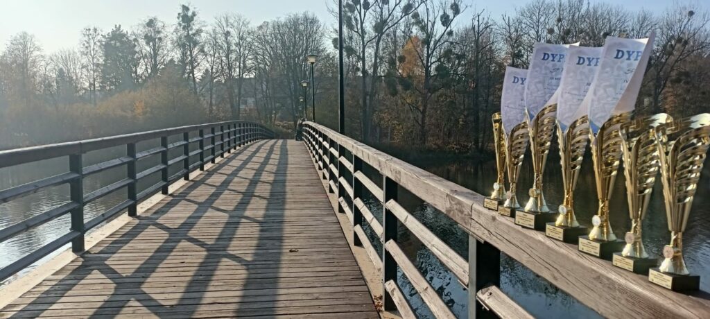 Na zdjęciu widoczny jest drewniany most nad wodą, biegnący w kierunku lasu, który widać w tle. Po prawej stronie mostu ustawione są złote puchary z włożonymi dyplomami, tak jak na poprzednich zdjęciach. Każdy puchar ma przy podstawie tabliczkę, a w kielichu znajdują się dyplomy informujące o osiągnięciach i zajętych miejscach przez szkołę SP 7. Most rzuca długie cienie na drewnianą podłogę, a w oddali widoczne są lampy oświetleniowe biegnące wzdłuż jego krawędzi. Tło wypełniają nagie, jesienne drzewa, co podkreśla chłodny, jesienny klimat zdjęcia. Cała scena jest oświetlona miękkim światłem, które tworzy spokojny, nostalgiczny nastrój.