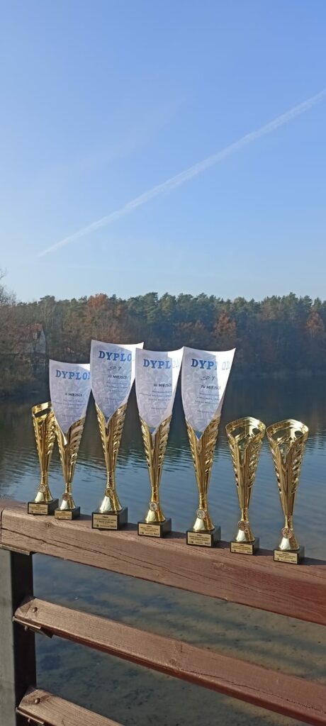 Na zdjęciu znajduje się sześć złotych pucharów ustawionych na drewnianej barierce, z widokiem na jezioro w tle. Trzy z pucharów mają włożone dyplomy, na których widać napisy „Dyplom” oraz informacje o zajętych miejscach (trzecie i czwarte miejsce) przez SP 7. Dyplomy są włożone w kielichy pucharów, co tworzy elegancką kompozycję. W tle widoczna jest tafla jeziora oraz las w jesiennych barwach, a nad nim rozciąga się jasne, bezchmurne niebo. Zdjęcie ma spokojny, jesienny nastrój, a kolory natury kontrastują z błyszczącymi pucharami na pierwszym planie.