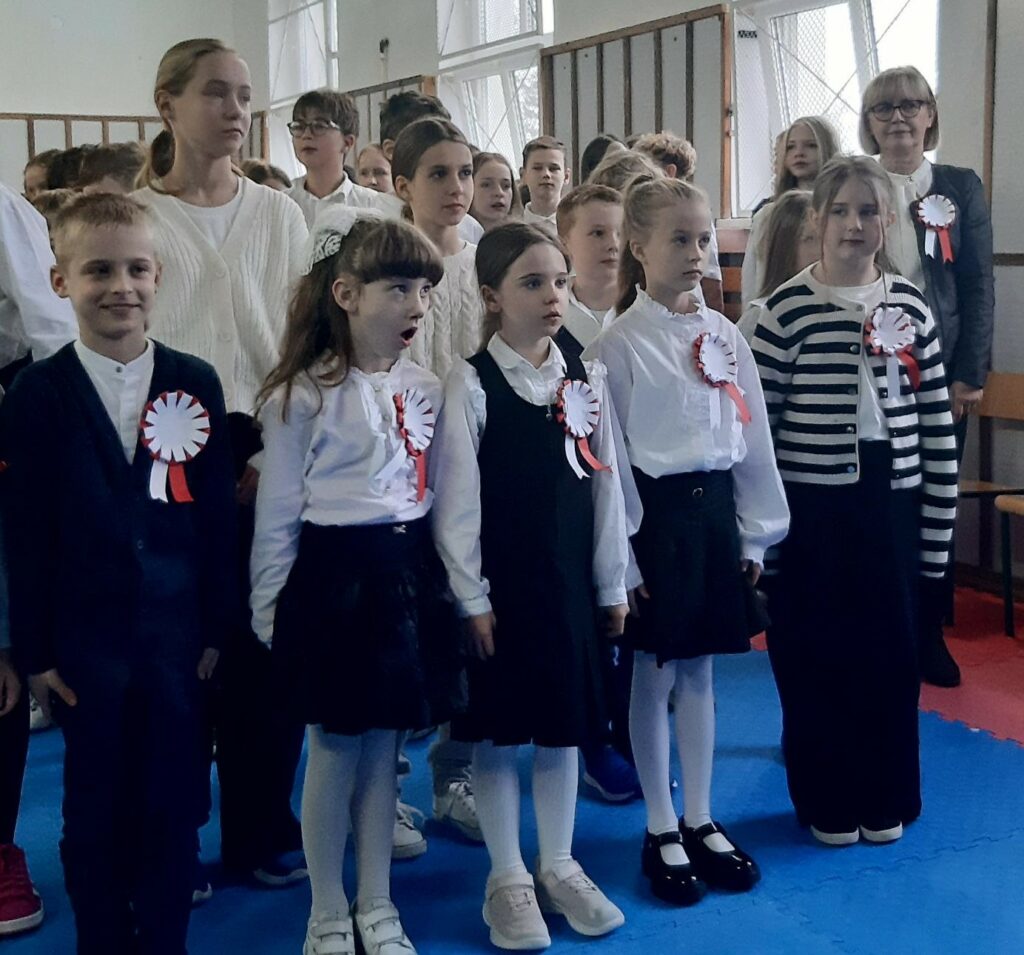 Na pierwszym planie liczna grupa dzieci stoi na baczność, w stroju galowym i przypiętym na piersi kotylionie.