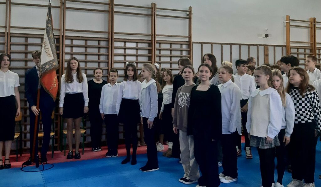 Grupa dzieci w pozycji na baczność i stroju galowym. W lewej części młodzież ze sztandarem szkoły. Wszyscy gotowi do rozpoczęcia wspólnego śpiewania “Mazurka Dąbrowskiego”