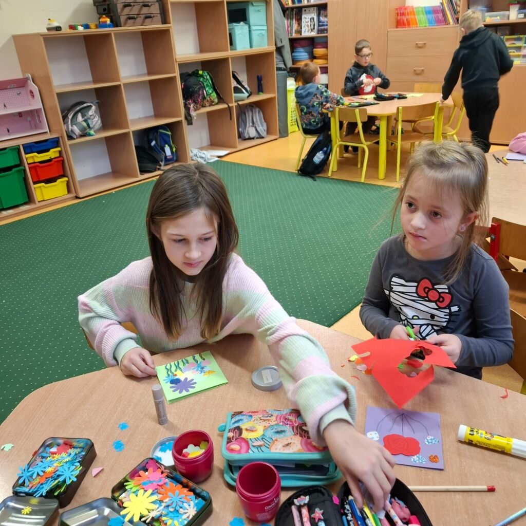 Zdjęcie przedstawia dwie dziewczynki siedzące przy sześciokątnym stole. Rysują one laurki. Na brązowym stole leżą kolorowe kartki, kolorowe kwiatki i inne papierowe ozdoby. W oddali widać inny stół i siedzące przy nim dzieci. Widać zielony dywan a w oddali brązowe szafki z kolorowymi zabawkami