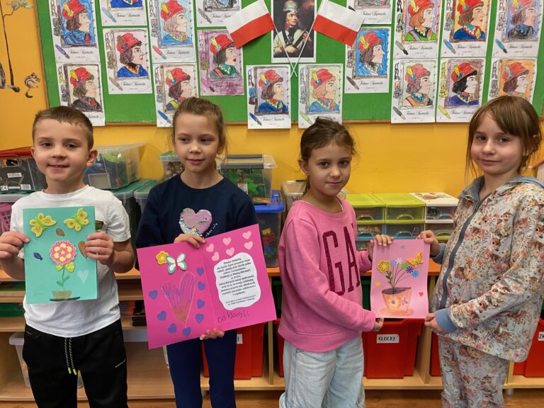 Na zdjęciu czworo dzieci przedstawia laurki, jakie wykonali dla seniorów. Dzieci ubrane są w kolorowe ubrania, w rękach trzymają kolorowe laurki. W tle widzimy gazetkę ścienną z pracami plastycznymi dzieci.