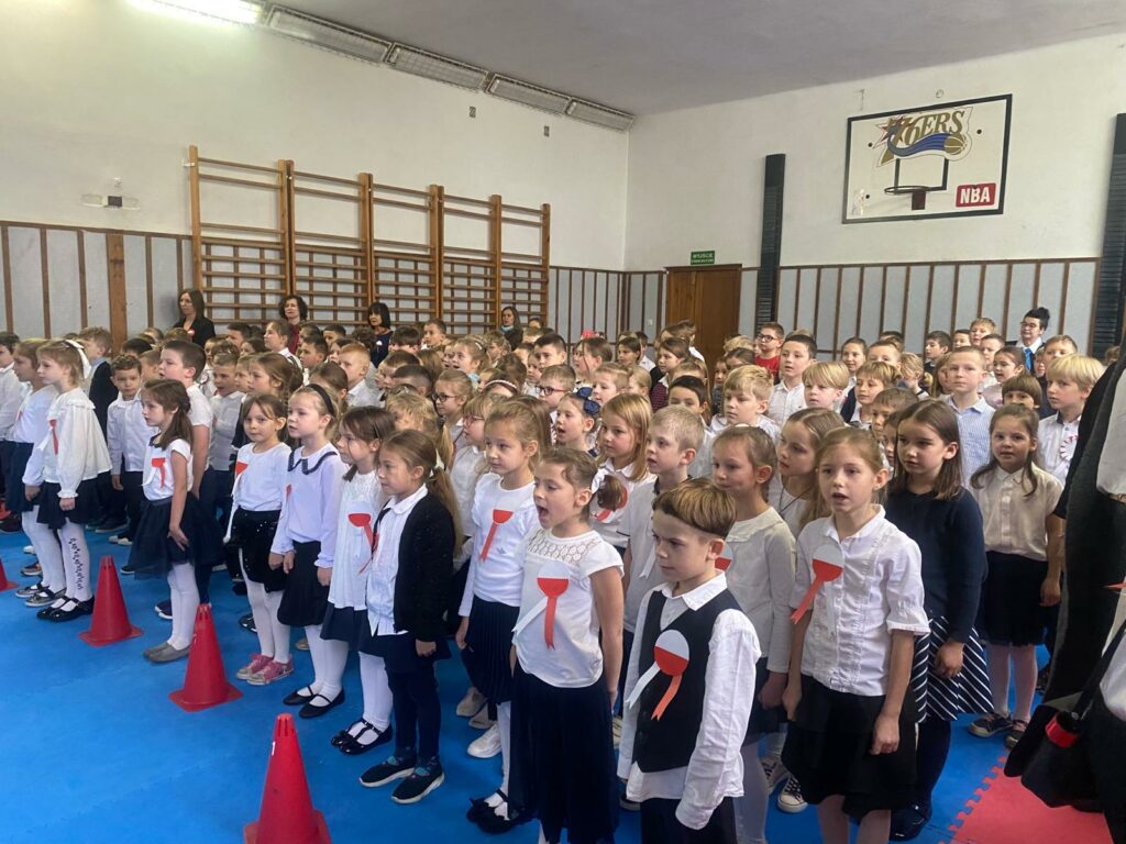 Kilkadziesięcioro dzieci stoi w kilku rzędach w pozycji na baczność. Wszystkie dzieci ubrane są na galowo, przy bluzkach mają wpięte kokardy narodowe. Przed dziećmi w pierwszym rzędzie ustawione są czerwone pachołki pachołki. Po lewej stronie zdjęcie widać drabinki , przed , którymi stoją cztery kobiety, jedna za drugą. u góry po prawej stronie zdjęcia widać kosz do gry w piłkę koszykową. Z tyłu po środku zdjęcia widoczne są drzwi.