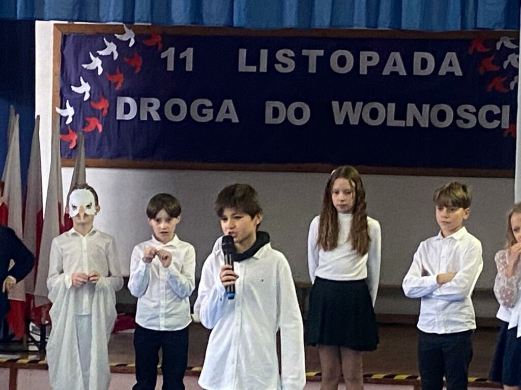 U góry na środku zdjęcia, tablica z napisem. Tło na tablicy jest granatowe i napis z białych liter: 11 listopada. Droga do wolności. Po obu stronach napisu, przypięte ptaki w kolorach biało- czerwonych lecące w górę. Na zdjęciu widoczne dzieci: od lewej stoi chłopiec w masce orła, następnie chłopiec ze złożonymi rękami, następnie chłopiec z mikrofonem, dziewczynka z rękoma do tyłu, chłopiec z założonymi rękoma. Po lewej stronie widać stojące flagi Polskie.