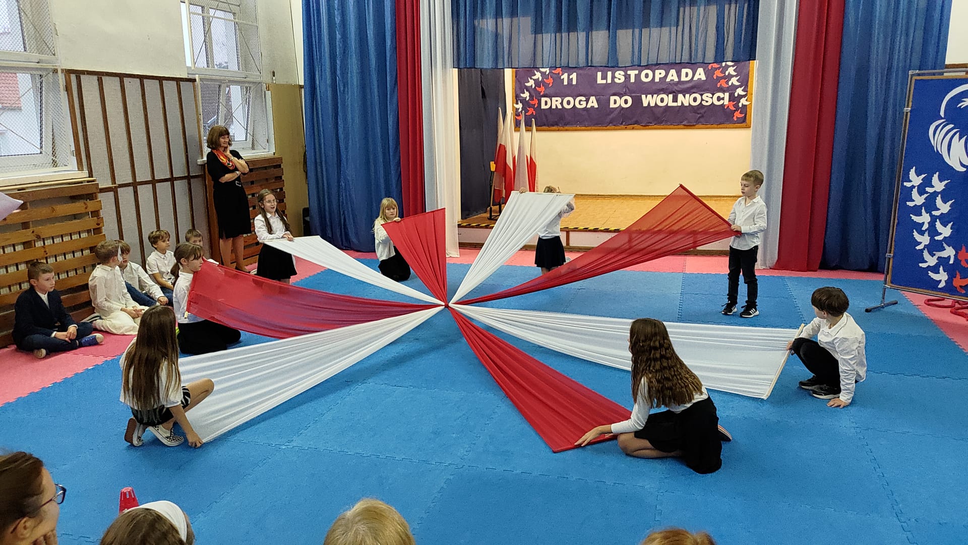 Widać scenę, u góry na środku zdjęcia, tablica z napisem. Tło na tablicy jest granatowe i napis z białych liter: 11 listopada. Droga do wolności. Po obu stronach napisu, przypięte ptaki w kolorach biało- czerwonych lecące w górę. Po prawej i lewej stronie zdjęcia zawieszony niebieski materiał i biało - czerwony. Po prawej stronie tablica z obrazkiem orła i ptakami biało- czerwonymi lecącymi w górę. Na środku zdjęcia ośmioro dzieci z szarfami , z których ułożona jest kokarda narodowa. Po lewej stronie siedzi pięciu chłopców ubranych na galowa i stojąca kobiety.