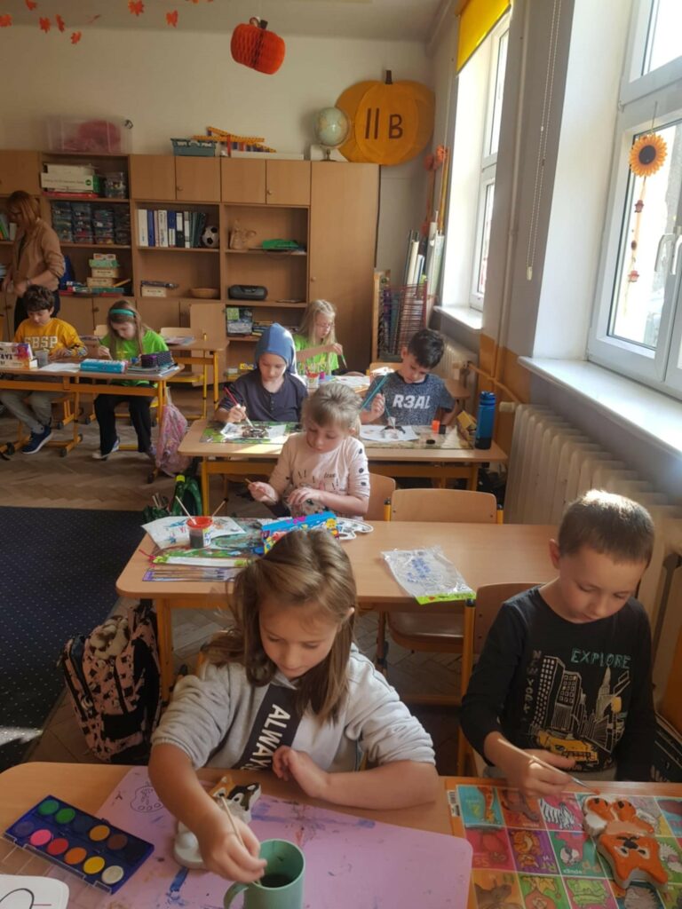 Na zdjęciu widać dzieci pracujące nad projektami plastycznymi w szkolnej klasie. Każde z nich jest pochylone nad swoim stolikiem, skupione na malowaniu i ozdabianiu różnych prac. Na ścianie wisi tablica korkowa z wyeksponowanymi rysunkami, a obok niej znajduje się półka z książkami i materiałami plastycznymi. W pomieszczeniu panuje ciepła, jesienna dekoracja, co widać po papierowych liściach i pomarańczowych ozdobach zawieszonych pod sufitem.
