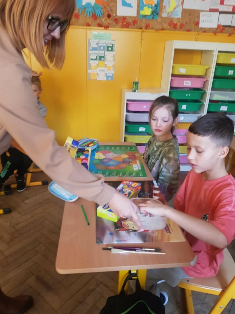 Na zdjęciu widzimy kobietę pochylającą się nad stolikiem, przy którym siedzi dwójka dzieci. Kobieta w okularach, ubrana w jasną bluzkę, pokazuje coś chłopcu w różowej koszulce, który trzyma pojemnik z masą plastyczną. Obok niego siedzi dziewczynka w zielonej koszulce, patrząc na zajęcia z zainteresowaniem. W tle widoczna jest tablica korkowa z dziecięcymi pracami plastycznymi oraz regał z kolorowymi pojemnikami na przybory szkolne.