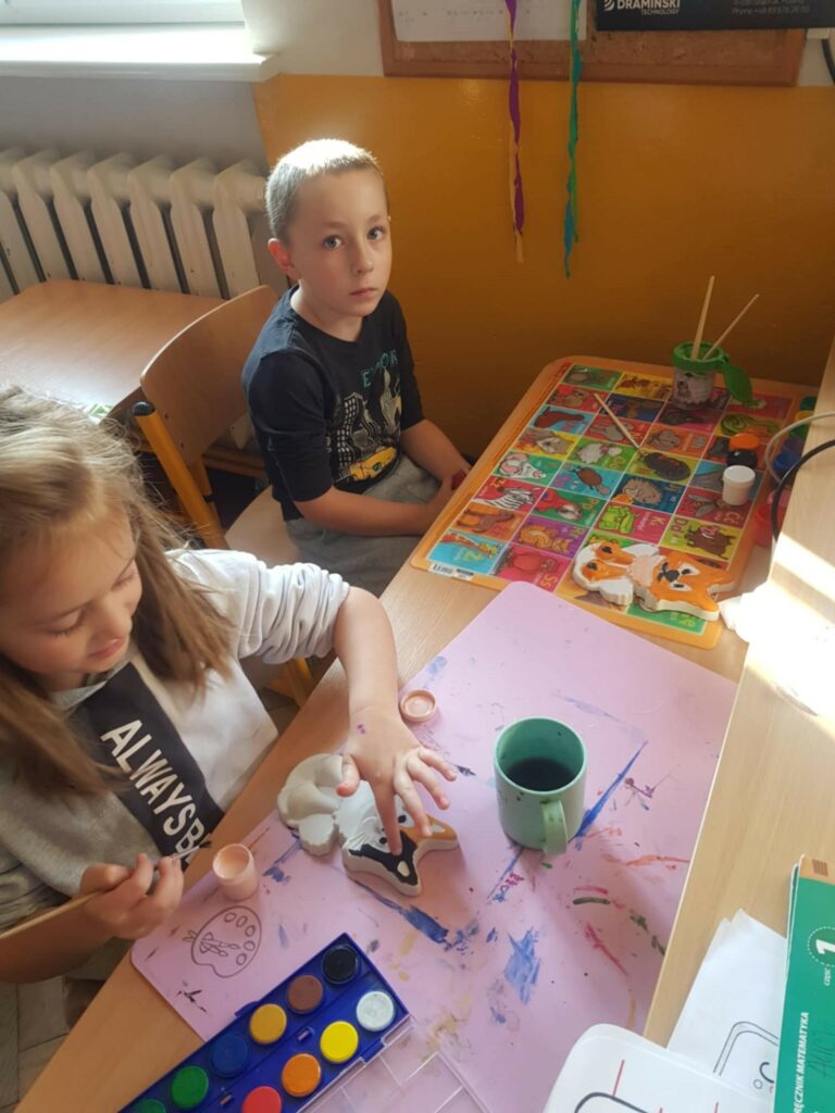 Na zdjęciu widzimy dwójkę dzieci siedzących przy stoliku w jasnym pomieszczeniu. Dziewczynka, ubrana w jasną bluzę z napisem "ALWAYS BE", zajmuje się malowaniem ceramicznej figurki, prawdopodobnie psa. Na stole przed nią leżą różne farby akwarelowe, kubek z wodą do płukania pędzli i różowy podkład zabezpieczający stół przed zabrudzeniem. Chłopiec obok, ubrany w ciemną koszulkę, patrzy prosto w obiektyw, sprawiając wrażenie, że jest mniej zaangażowany w malowanie. Na stole widać też kolorową podkładkę z obrazkami. W tle znajduje się kaloryfer oraz tablica korkowa na ścianie z żółtym paskiem na dole.