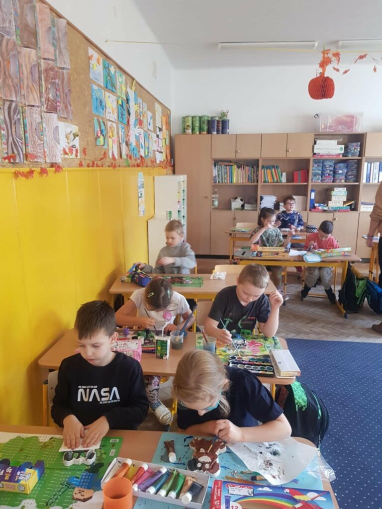 Na zdjęciu widać dzieci pracujące nad projektami plastycznymi w szkolnej klasie. Każde z nich jest pochylone nad swoim stolikiem, skupione na malowaniu i ozdabianiu różnych prac. Na ścianie wisi tablica korkowa z wyeksponowanymi rysunkami, a obok niej znajduje się półka z książkami i materiałami plastycznymi. W pomieszczeniu panuje ciepła, jesienna dekoracja, co widać po papierowych liściach i pomarańczowych ozdobach zawieszonych pod sufitem.