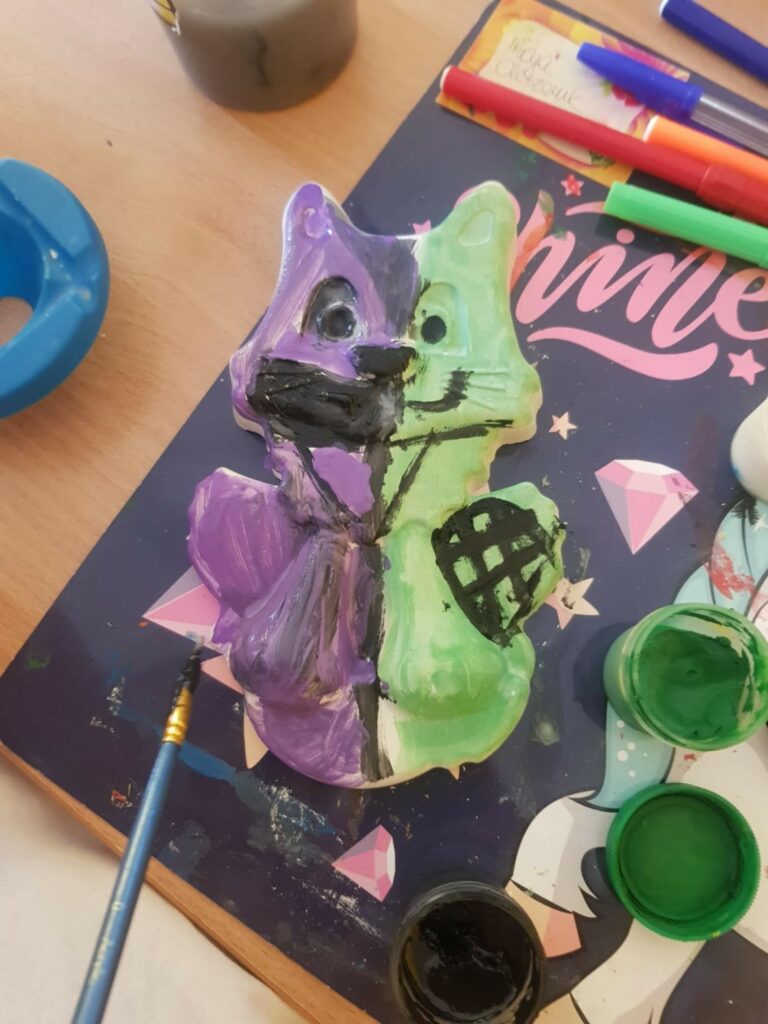 Na stoliku leży podkładka, a na niej odlew gipsowy w kształcie kota. Odlew został pomalowany różnymi kolorami: przeważa fiolet i zieleń. Obok stoją otwarte pojemniczki z farbą.