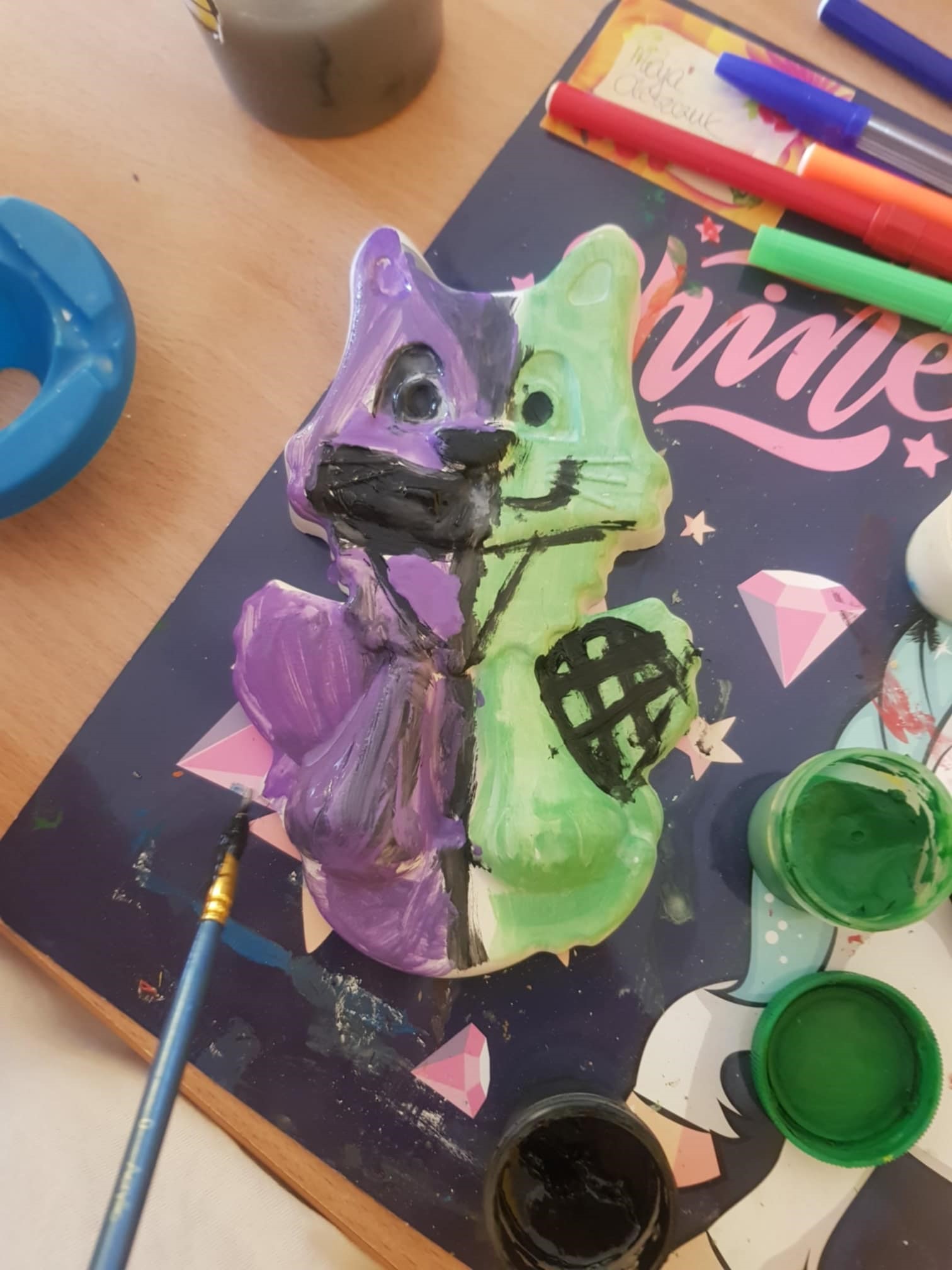 Na stoliku leży podkładka, a na niej odlew gipsowy w kształcie kota. Odlew został pomalowany różnymi kolorami: przeważa fiolet i zieleń. Obok stoją otwarte pojemniczki z farbą.