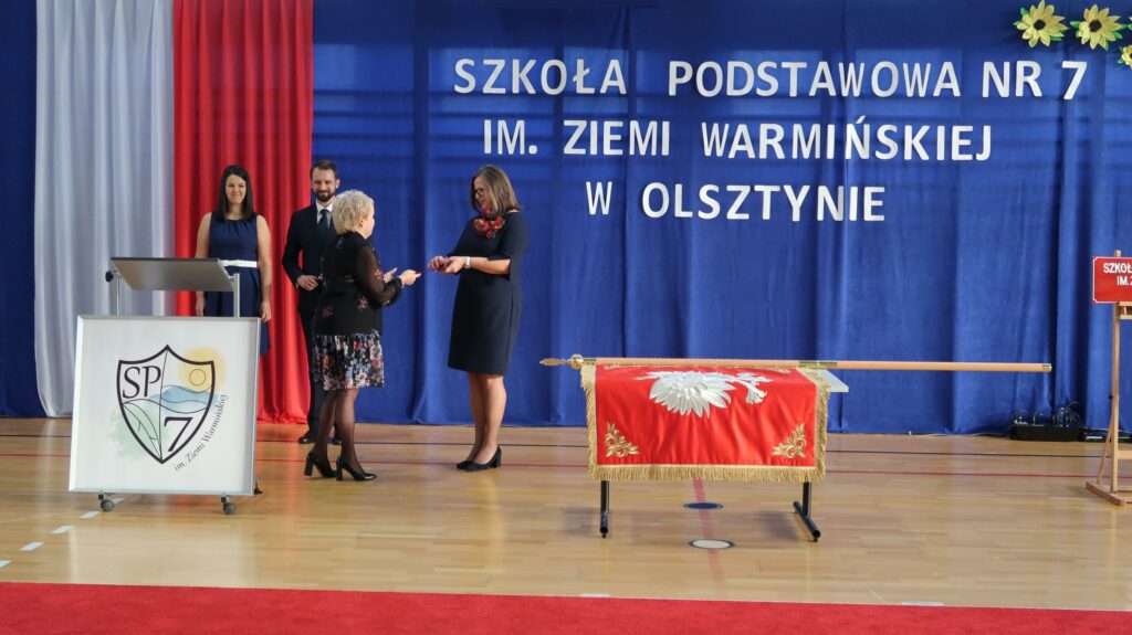Zdjęcie przedstawia uroczystość w Szkole Podstawowej nr 7 im. Ziemi Warmińskiej w Olsztynie, która odbywa się w sali gimnastycznej. Na środku, na tle niebieskiej dekoracji, kobieta w krótkich blond włosach przekazuje dokument kobiecie w granatowej sukience. Obok nich, po lewej stronie, stoi mównica z logo szkoły oraz dwie osoby – kobieta w granatowej sukience trzymająca mikrofon i mężczyzna w garniturze. Po prawej stronie, na stole leży sztandar szkolny, ozdobiony czerwonym materiałem z haftem oraz złotymi frędzlami. Tło zdjęcia stanowi przyczepiony napis z nazwą szkoły oraz zestaw żółtych kwiatów przypominających słoneczniki. Po lewej stronie flaga Polski.