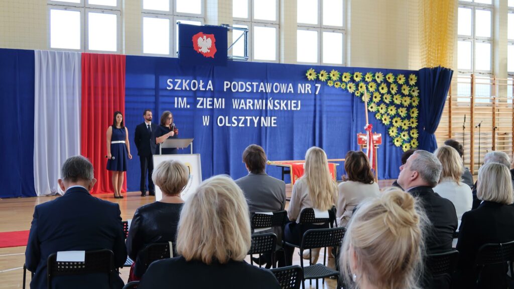 Zdjęcie przedstawia uroczystość w Szkole Podstawowej nr 7 im. Ziemi Warmińskiej w Olsztynie, która odbywa się w sali gimnastycznej. Na niebieskim tle, przy mównicy stoi kobieta, która wygłasza mowę. Obok niej, po lewej stronie, znajdują się dwie osoby: kobieta z mikrofonem i mężczyzna w garniturze. Nad nimi widnieje napis z nazwą szkoły oraz godło Polski. Po prawej stronie od napisu widać żółte słoneczniki. Na pierwszym planie widać siedzących na krzesłach zaproszonych gości.