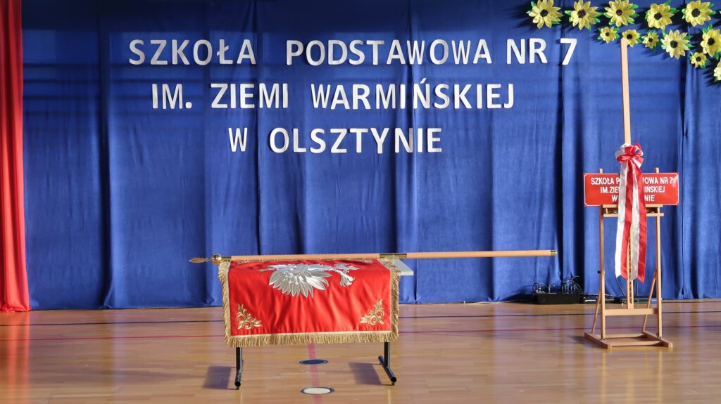 Zdjęcie przedstawia scenę udekorowaną na uroczystość nadania imienia w Szkole Podstawowej nr 7 im. Ziemi Warmińskiej w Olsztynie. W centrum, na stole, znajduje się sztandar szkoły ozdobiony czerwonym materiałem z haftem oraz złotymi frędzlami. W tle jest niebieska kotara, na której znajduje się biały napis: „SZKOŁA PODSTAWOWA NR 7 IM. ZIEMI WARMIŃSKIEJ W OLSZTYNIE”. Po prawej stronie znajduje się czerwona tablica z nazwą szkoły, przyozdobiona dużą biało-czerwona kokardą, ustawiona na drewnianym stojaku. Dekorację uzupełniają żółte kwiaty przypominające słoneczniki.