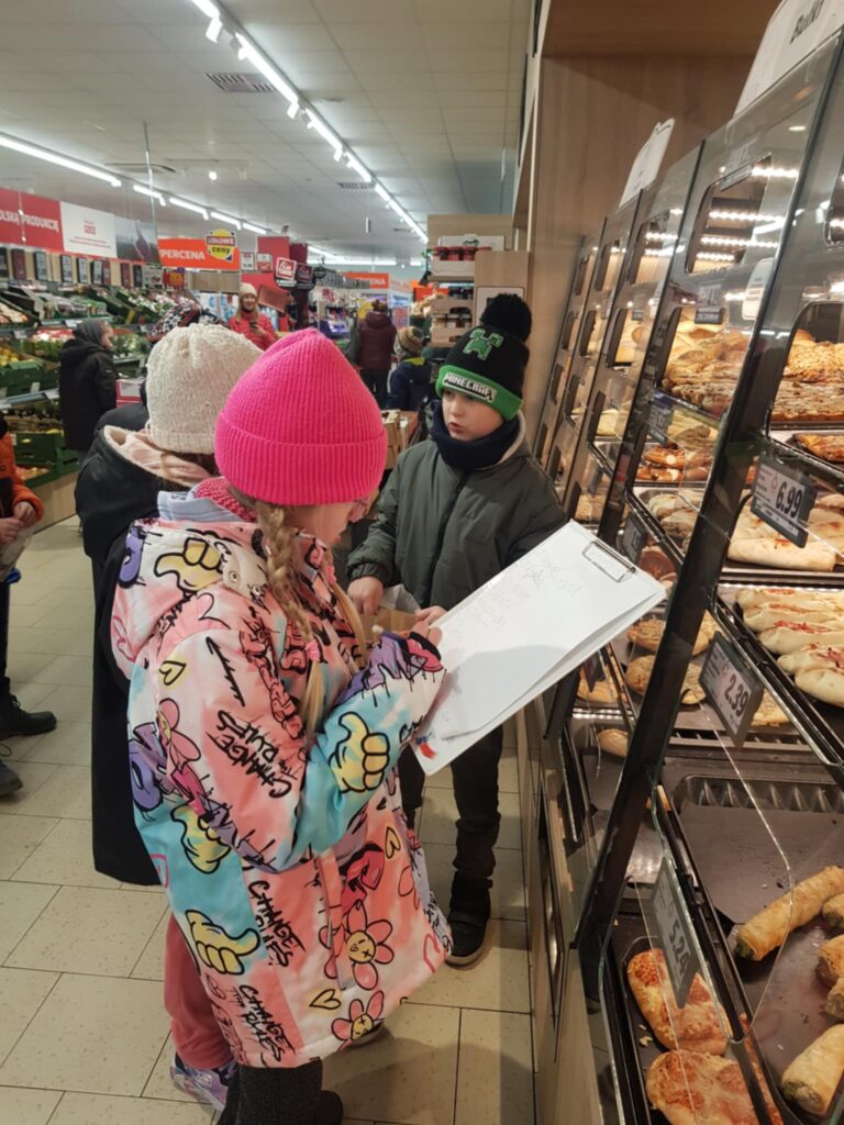 W sklepie, troje dzieci stoi przy regałąch z pieczywem. Zapisują ceny na karcie pracy. W tle widać regały i kupujacych.