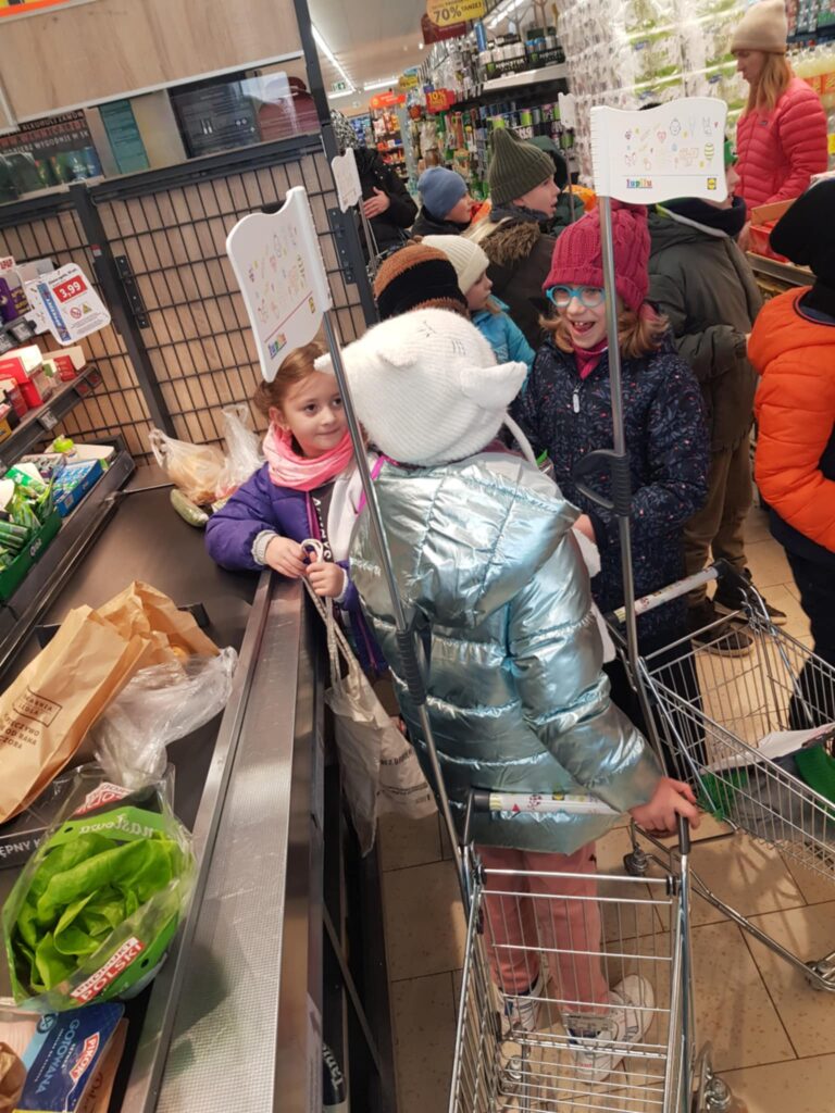 W sklepie, kilkoro dzieci stoi przy kasie. Wykładają produkty na taśmę. Dwoje dzieci ma przed soba małe wózki na zakupy. W tle widać regały i kupujacych.