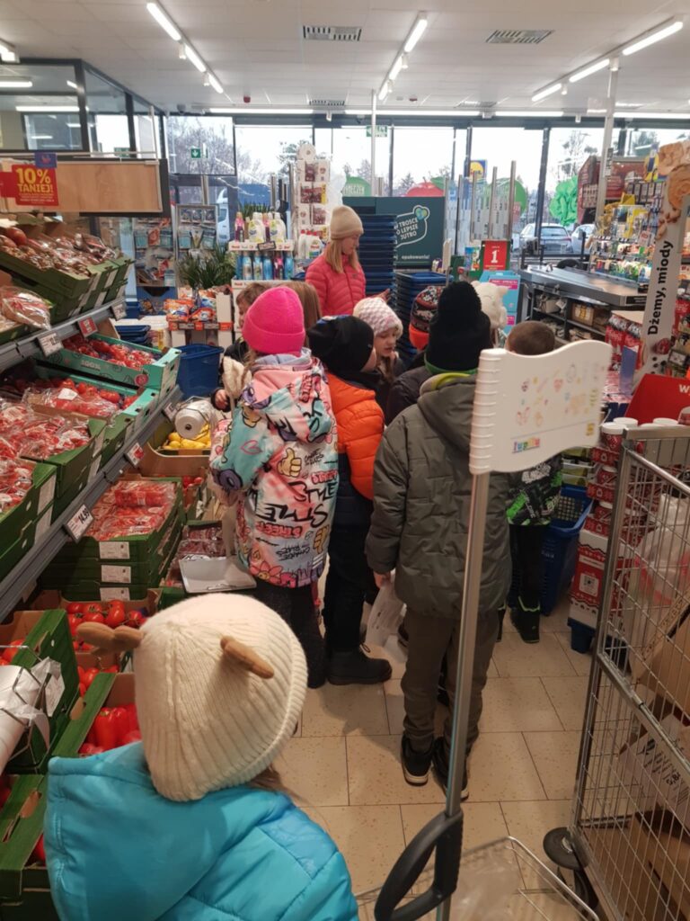 W sklepie, między regałami, stoi grupa dzieci, odwrócona tyłem do obiektywu. Wybierają warzywa, aby je kupić. Jedno z dzieci pcha przed sobą mały wózek na zakupy. W tle widać regały sklepowe z towarami.