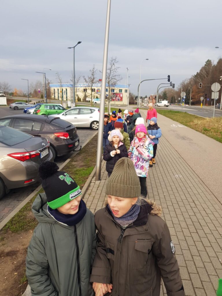 Klasa idzie w parach po chodniku. Dzieci ubrane sa w kurtki, czapki i szaliki. Po lewej stronie widać zaparkowane samochody, po prawej- pas zieleni, ulice i jadace samochody . W pierwszym planie widać dwóch chłopców.