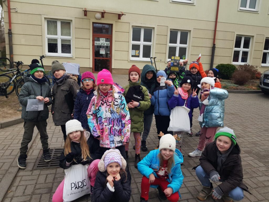 Grupa dzieci stoi przed szkołą. Przed nimi kilkoro dzieci kuca. Wszyscy są ciepło ubrani. Niektóre dzieci trzymają torby z zakupami, a niektóre woreczki z pieniędzmi i paragonami. Dzieci są uśmiechnięte.