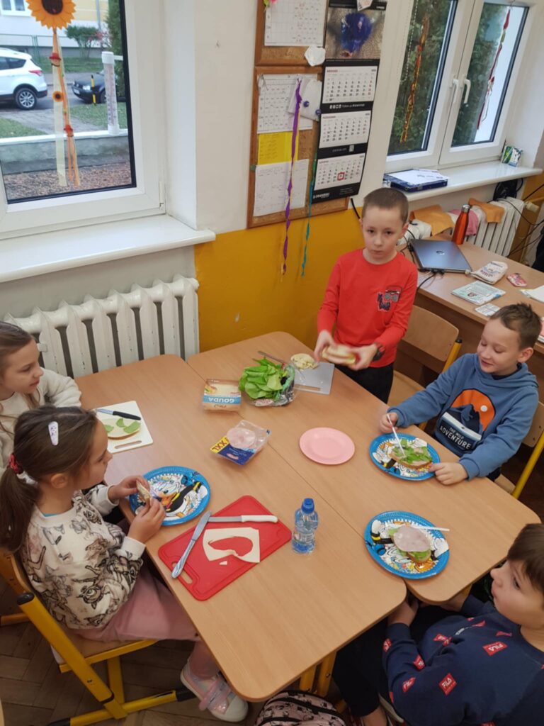 W klasie, w centralnym miejscu, widać dwa złączone stoły. Wokół nich pięcioro dzieci siedzi na krzesełkach. Przygotowują kanapki. na stołach leżą talerze papierowe, serwetki i produkty spożywcze na kanapki. W tle widać okno z widokiem i tablicę z ogłoszeniami.