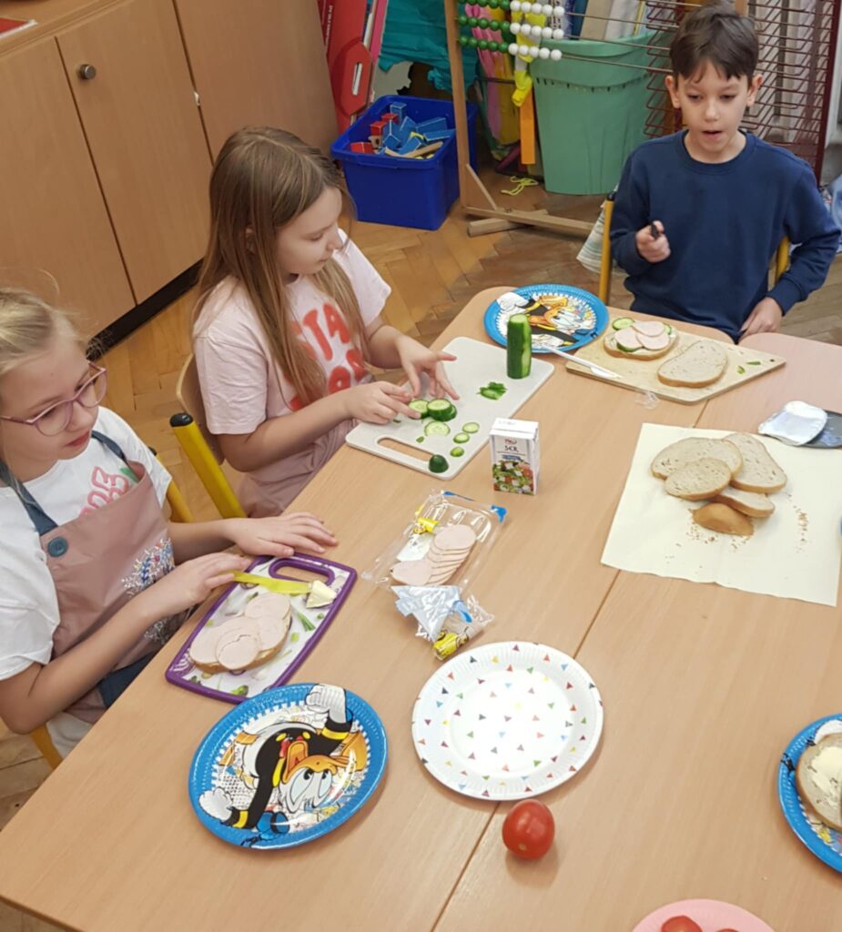 W klasie, w centralnym miejscu, widać dwa złączone stoły. Wokół nich dzieci siedzi na krzesełkach. Przygotowują kanapki. na stołach leżą talerze papierowe, serwetki i produkty spożywcze na kanapki. W tle widać szafę i pojemniki z klockami.