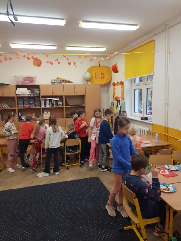 Zdjęcie przedstawia pomieszczenie klasowe. Widok z góry przedstawia kilka złączonych ławek i dzieci. Niektóre siedzą i robią kanapki, niektóre stoją. Na środku klasy leży wykładzina. W tle widać szafki.