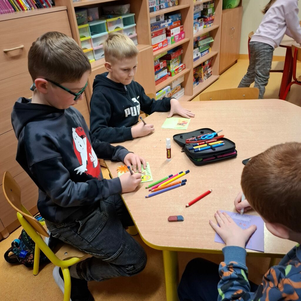 Zdjęcie przedstawia trójkę chłopców siedzących przy sześciokątnym stole, którzy rysują laurki. Stół jest brązowego koloru, na stole leżą kolorowe kartki, piórnik, kolorowe kredki. W tle widać brązowe szafki z kolorowymi zabawkami.