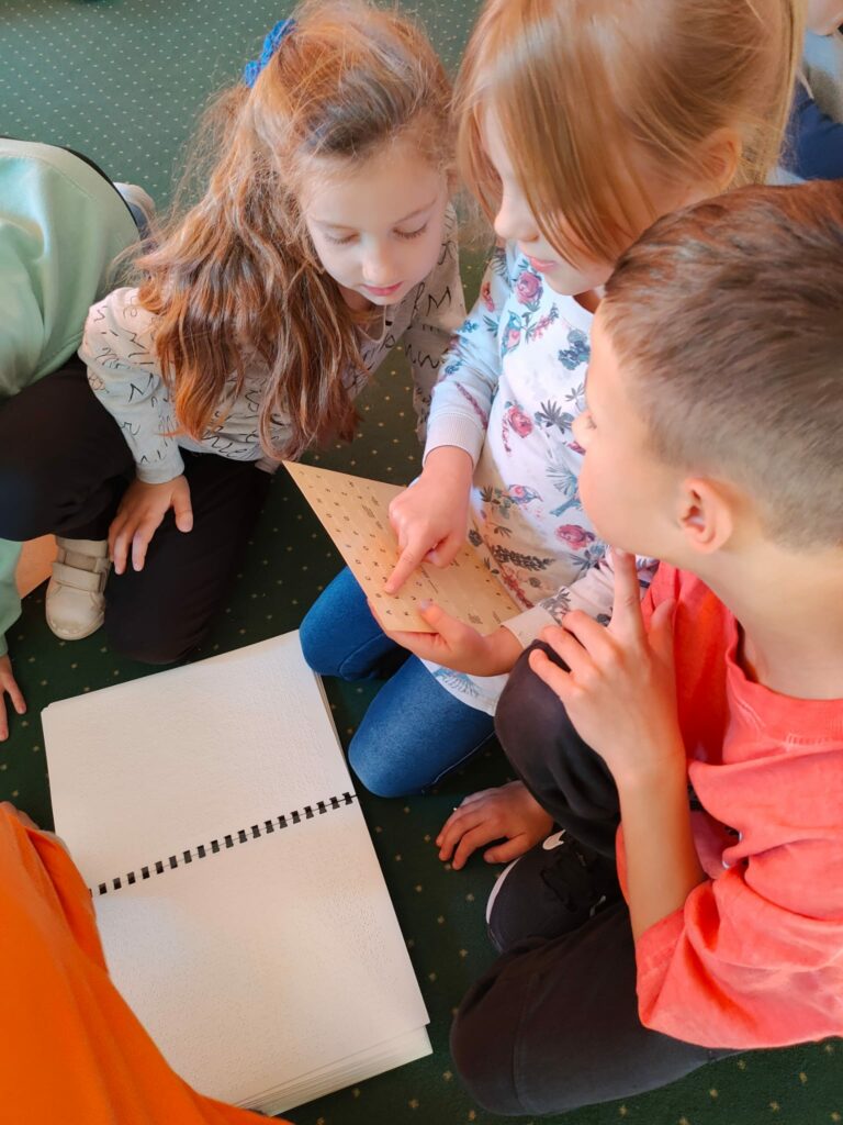 Na zdjęciu troje dzieci pochyla się nad kartką z literami. Siedząca po środku dziewczynka wskazuje palcem coś na kartce. Ta dziewczynka ubrana jest w białą bluzkę i granatowe dżinsy. Bluzka ma kwiatowy wzór. Chłopiec siedzący po prawej stronie ubrany jest w różową koszulkę. Trzecie dziecko to dziewczynka z długimi włosami. Dzieci siedzą na zielonym dywanie. Na dywanie lezy biały, otwarty zeszyt.