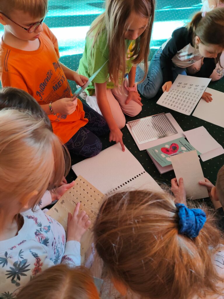 Zdjęcie przedstawia grupę pięciorga dzieci siedzących na zielonym dywanie. Siedzą wokół rozłożonych na dywanie kartek i zeszytów oraz kolorowej książki. Jeden z chłopców trzyma w ręku kartkę i wskazuje palcem literę . Skierowany w naszą stronę tekst kartki pokazuje że jest to alfabet Braille’a. Chłopiec w pomarańczowej koszulce trzyma dwoma rękoma kartkę i patrzy na nią. Tło za dziećmi jest koloru seledynowego.