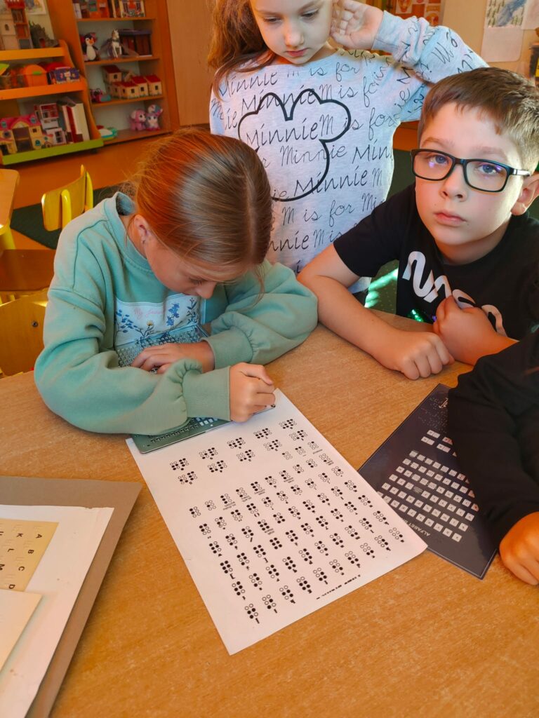 Na zdjęciu widać trójkę dzieci pochylających się nad brązowym blatem stołu. Chłopiec i dziewczynka siedzą przy stole. Inna dziewczynka stoi za nimi i w skupieniu przygląda się co dzieje się na stole. Na stole leżą kartki z alfabetem Braille’a. Siedząca za stołem dziewczynka pochyla się i coś pisze na lezącej na stole białej kartce .