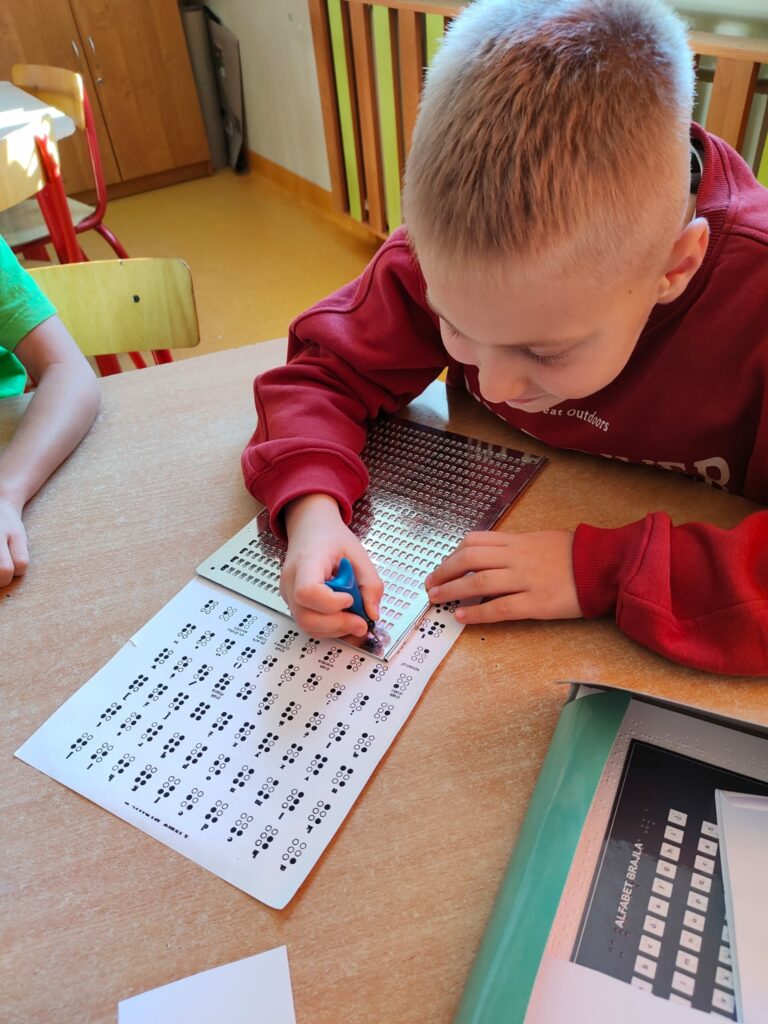 Na zdjęciu widać jak chłopiec w bordowej bluzie siedzi przy stole. Pochyla się nad kartką z alfabetem Braille’a. Pisze litery przy użyciu szablonu. W tle widać krzesło, podłogę , brązowo – zieloną obudową do kaloryfera oraz kawałek brązowej szafki.