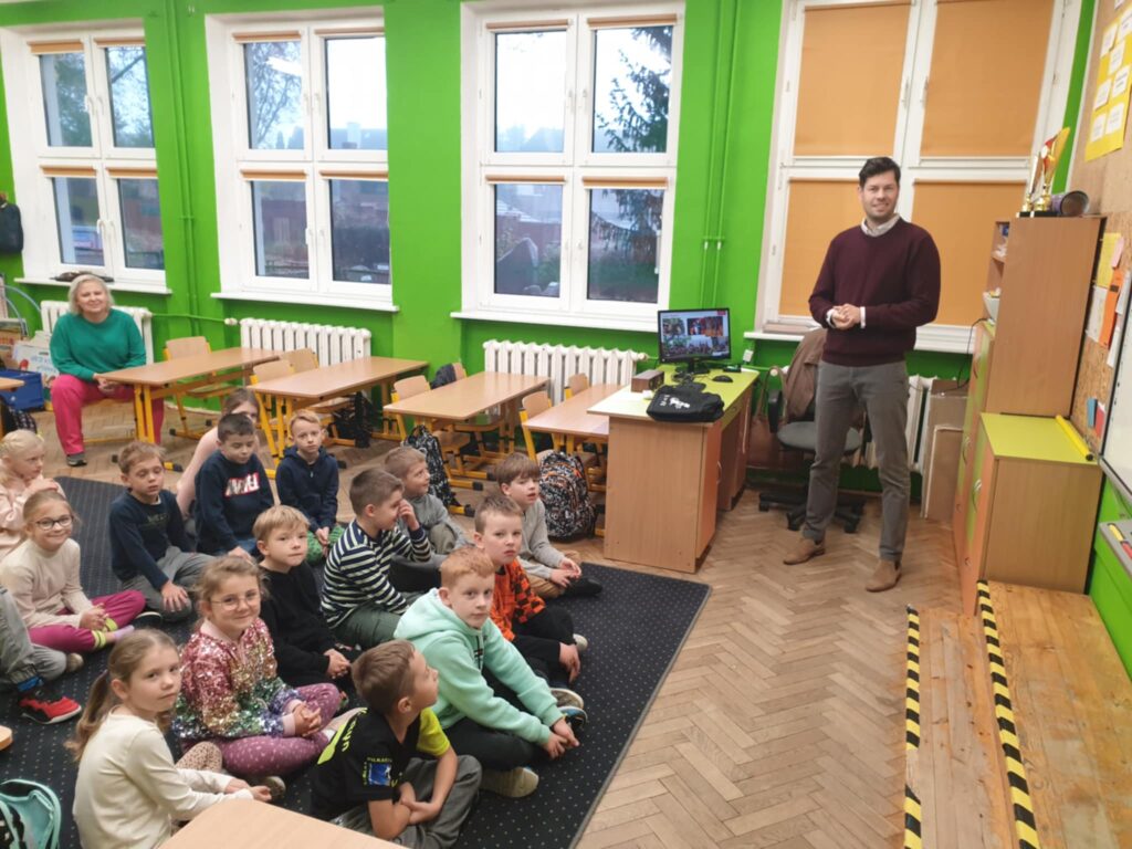 Na zdjęciu widać lekcję w klasie, podczas której mężczyzna prezentuje dzieciom informacje na tablicy multimedialnej. Dzieci siedzą na dywanie z przodu klasy, zwrócone twarzą do mężczyzny i tablicy, uważnie obserwując. Klasa jest kolorowa, z zielonymi i pomarańczowymi ścianami oraz zasłoniętymi żaluzjami w oknach, co nadaje przytulny nastrój. Na tablicy wyświetlane są różne zdjęcia i tekst, prawdopodobnie związane z tematem prezentacji, co wzbudza zainteresowanie uczniów. Atmosfera wydaje się spokojna, a dzieci są skupione na przekazywanych informacjach.