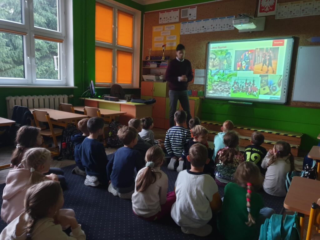 Na zdjęciu widać klasę szkolną, w której dzieci siedzą na dywanie przed tablicą i uważnie słuchają mężczyzny. W tle znajduje się kobieta siedząca na krześle, która jest nauczycielką. Po prawej stronie mężczyzna, prowadzący zajęcia, stoi przy biurku z komputerem i plecakiem na blacie. Klasa jest pomalowana na intensywny zielony kolor, co dodaje wnętrzu energii. Na ścianie obok tablicy widać różne pomoce dydaktyczne, które mogą być wykorzystywane podczas lekcji.