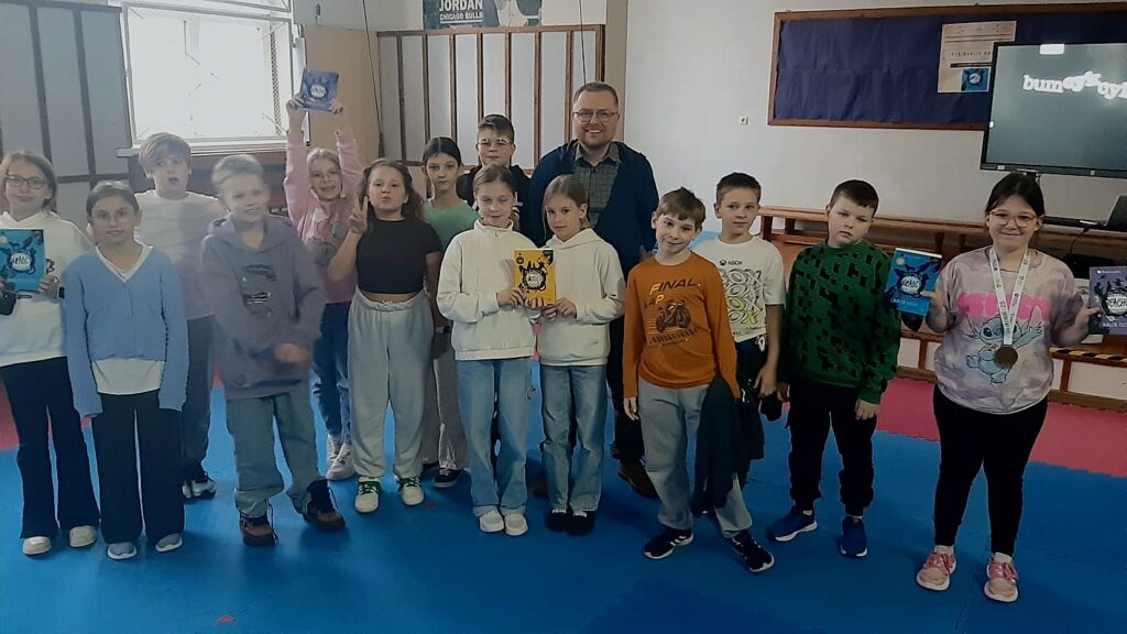 Zdjęcie przedstawia grupę dzieci niektóre mają w dłoniach książki. Między nimi stoi uśmiechnięty mężczyzna w okularach. Za grupą widać fragment multimedialnego telewizora, tablicę na ścianie z granatowym tłem na której widać powieszony plakat .