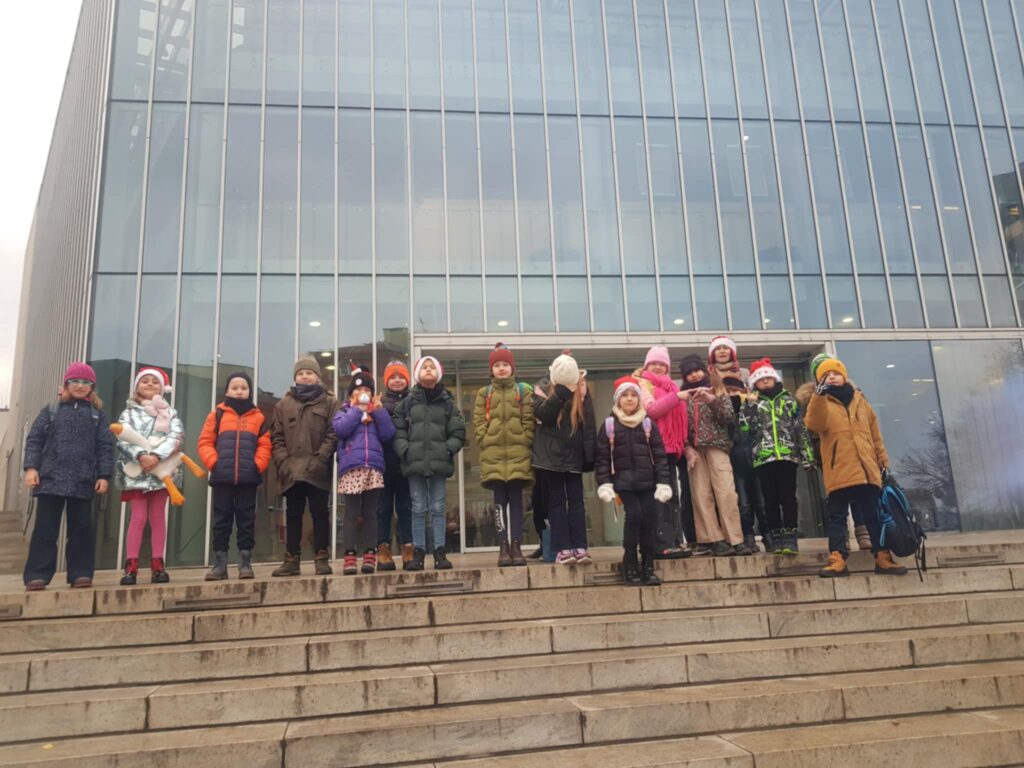 Przed wielkim oszklonym budynkiem, stoi na schodach grupa dzieci z nauczycielką, Jest to filharmonia. Dzieci ubrane są w kurtki i czapki.