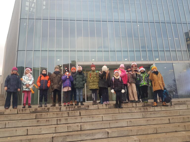 Przed wielkim oszklonym budynkiem, stoi na schodach grupa dzieci z nauczycielką, Jest to filharmonia. Dzieci ubrane są w kurtki i czapki.