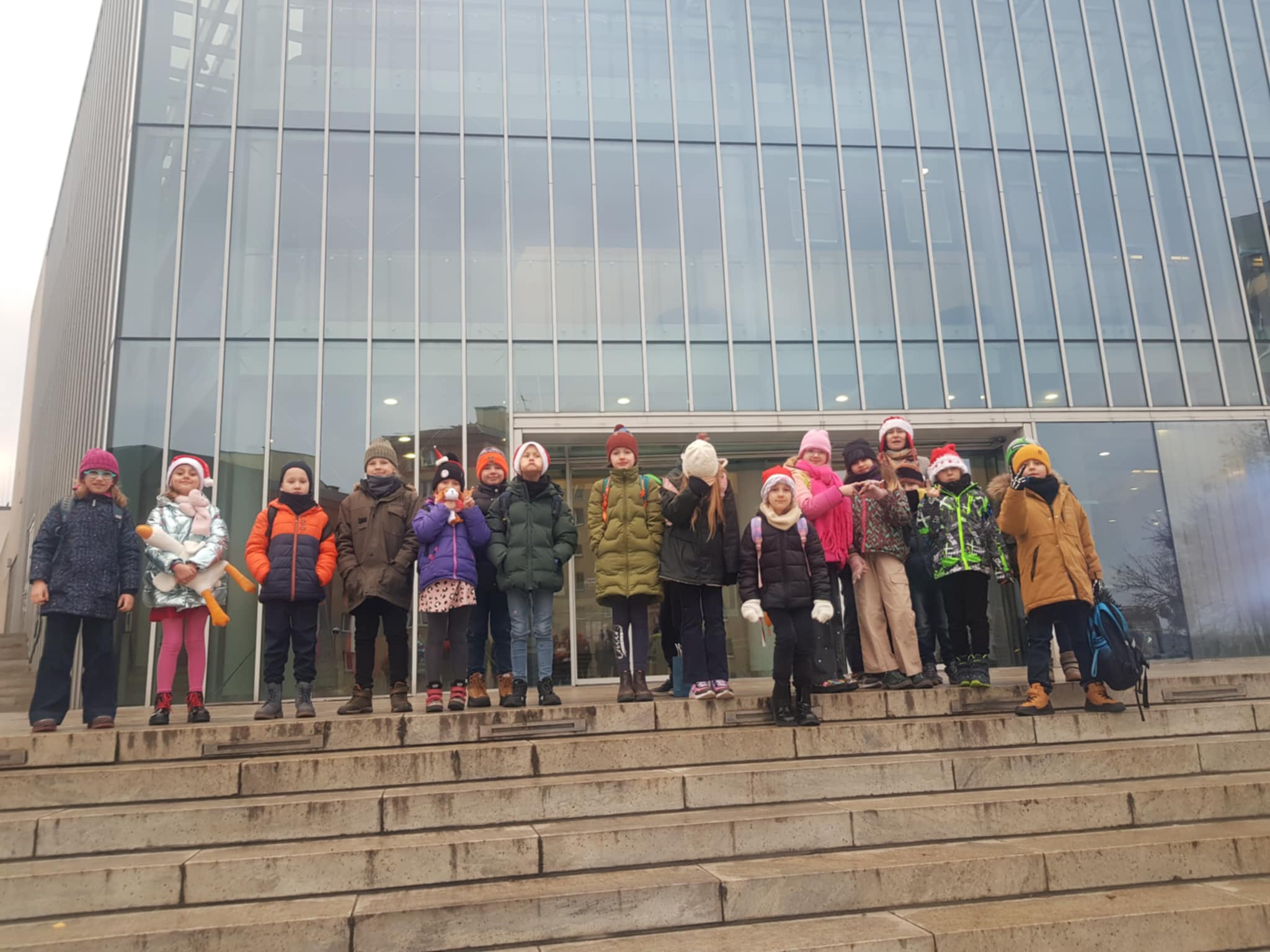 Przed wielkim oszklonym budynkiem, stoi na schodach grupa dzieci z nauczycielką, Jest to filharmonia. Dzieci ubrane są w kurtki i czapki.