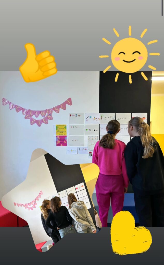 Na zdjęciu widać grupę uczennic oglądających wystawę na ścianie, na której znajdują się prace – rebusy, związane z Dniem Życzliwości. Ściana ozdobiona jest różowym napisem „Dzień Życzliwości”. Kolorowe rysunki i plakaty przyciągają uwagę. Zdjęcie uzupełniają emoji słońca, serca i uniesionego kciuka.