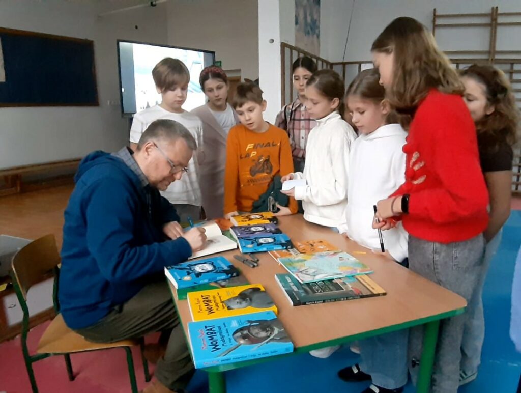 Mężczyzna w okularach i granatowej bluzie siedzi przy stoliku na którym rozłożone są książki, pisaki. Mężczyzna podpisuje książkę. Wokół stolika stoją dzieci przyglądające się czynności. Za dziećmi widać częściowo ekran, drabinki gimnastyczne.