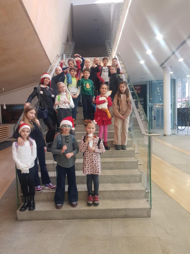 Grupa dzieci stoi na schodach wewnątrz budynku. Dzieci mają ubrania z elementami świątecznymi. Hol jest przestronny.