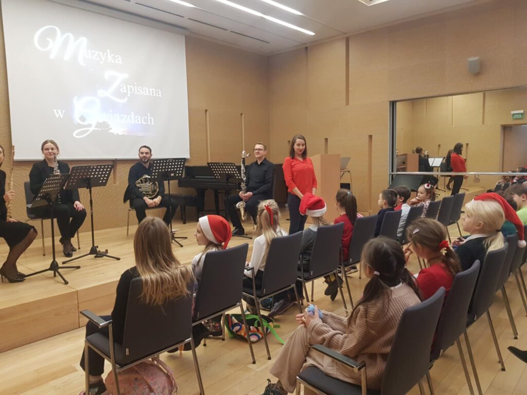 zdjęcie przedstawia niewielką salę koncertową. Na scenie widać czterech muzyków, obok stoi kobieta prowadzaca spotkanie. Na widowni siedzi kilkanaścioro dzieci. Na dużym ekranie za nimi wyświetlany jest napis „Muzyka zapisana w gwiazdach”.