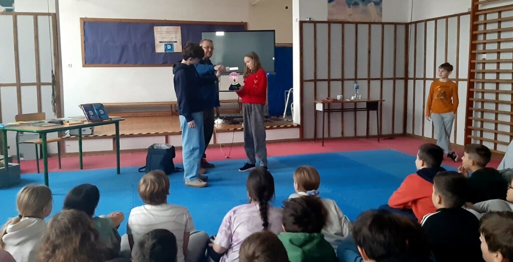 Mężczyzna przeprowadza doświadczenie z udziałem dzieci, które aktywnie uczestniczą w pokazie. Dzieci trzymają rekwizyty naukowe, dziewczynka kulę plazmową, chłopiec trzyma w dłoni dużą żarówkę mężczyzna pomaga mu przybliżyć ją do kuli. Z boku w oddaleniu stoi chłopiec koło drabinek gimnastycznych. Za grupą z doświadczeniami widać stolik z książkami, ekran. Przed grupą siedzą widzowie.